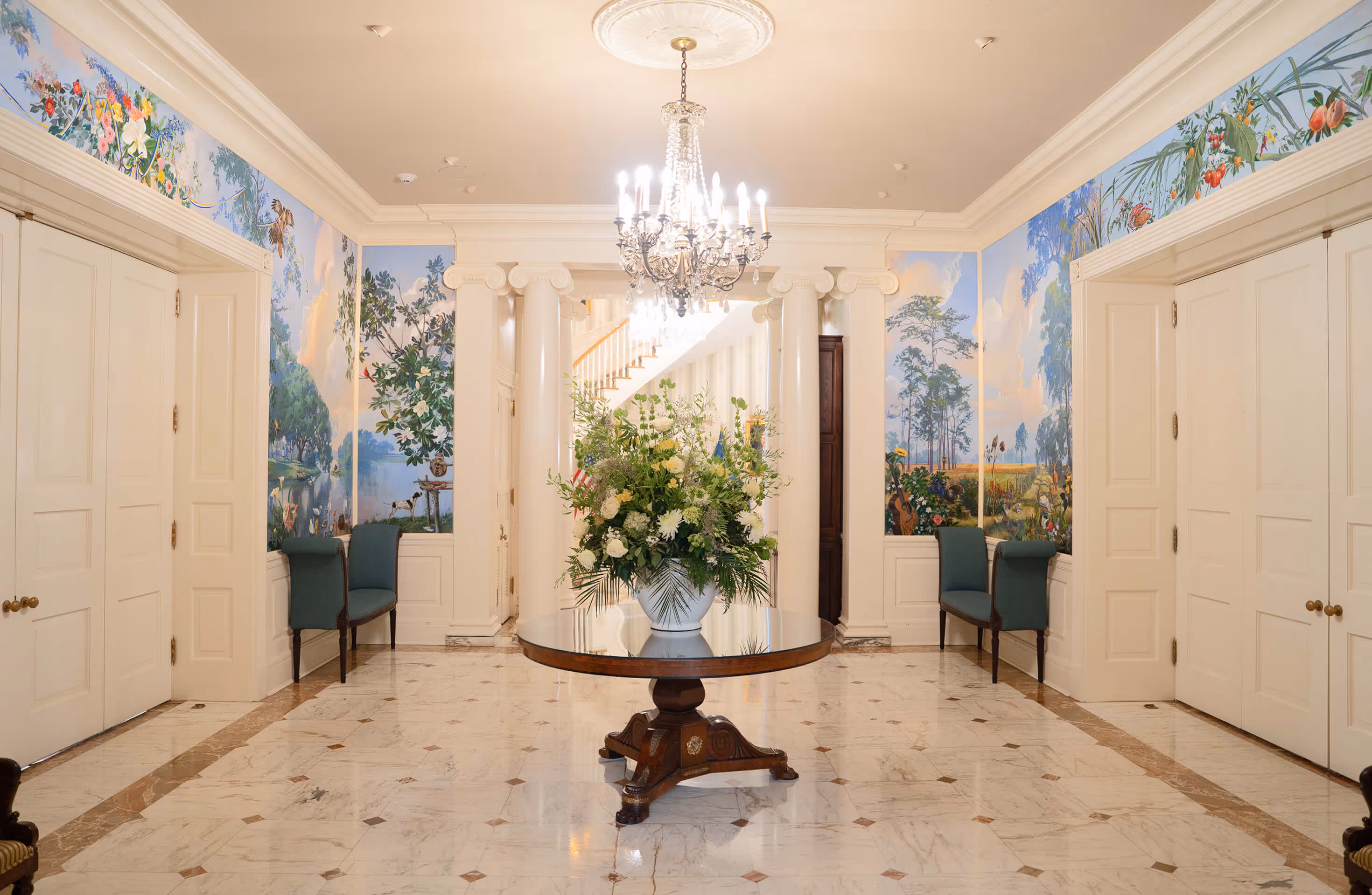 Elegant foyer with a round wooden table holding a large floral arrangement, crystal chandelier, marble floor, and mural-covered walls with chairs.