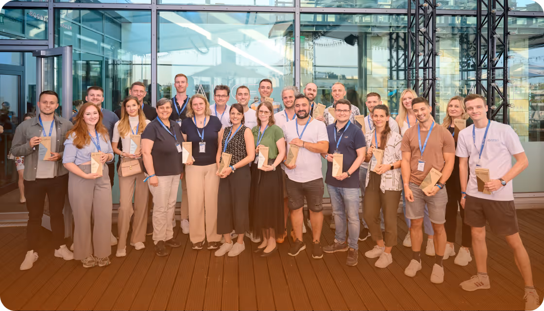 Gruppe von Männern und Frauen mit Ausweisen und braunen Geschenktüten, die auf einer Holzterrasse vor einem Glasgebäude posieren.