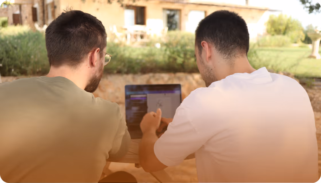 Zwei Männer sitzen draußen an einem Tisch und schauen gemeinsam auf einen Laptopbildschirm.