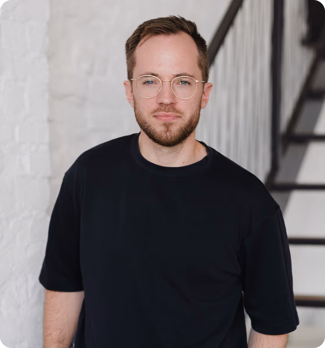 Porträt eines Mannes mit Bart und Brille, der ein schwarzes T-Shirt trägt und vor einer weißen Wand neben einer Treppe steht.
