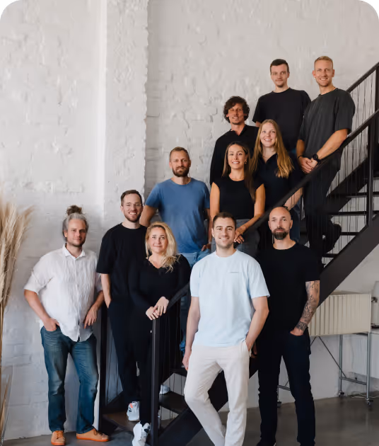Gruppenfoto von elf lächelnden Personen, die auf und neben einer Treppe in einem hellen Raum mit weißer Backsteinwand stehen.