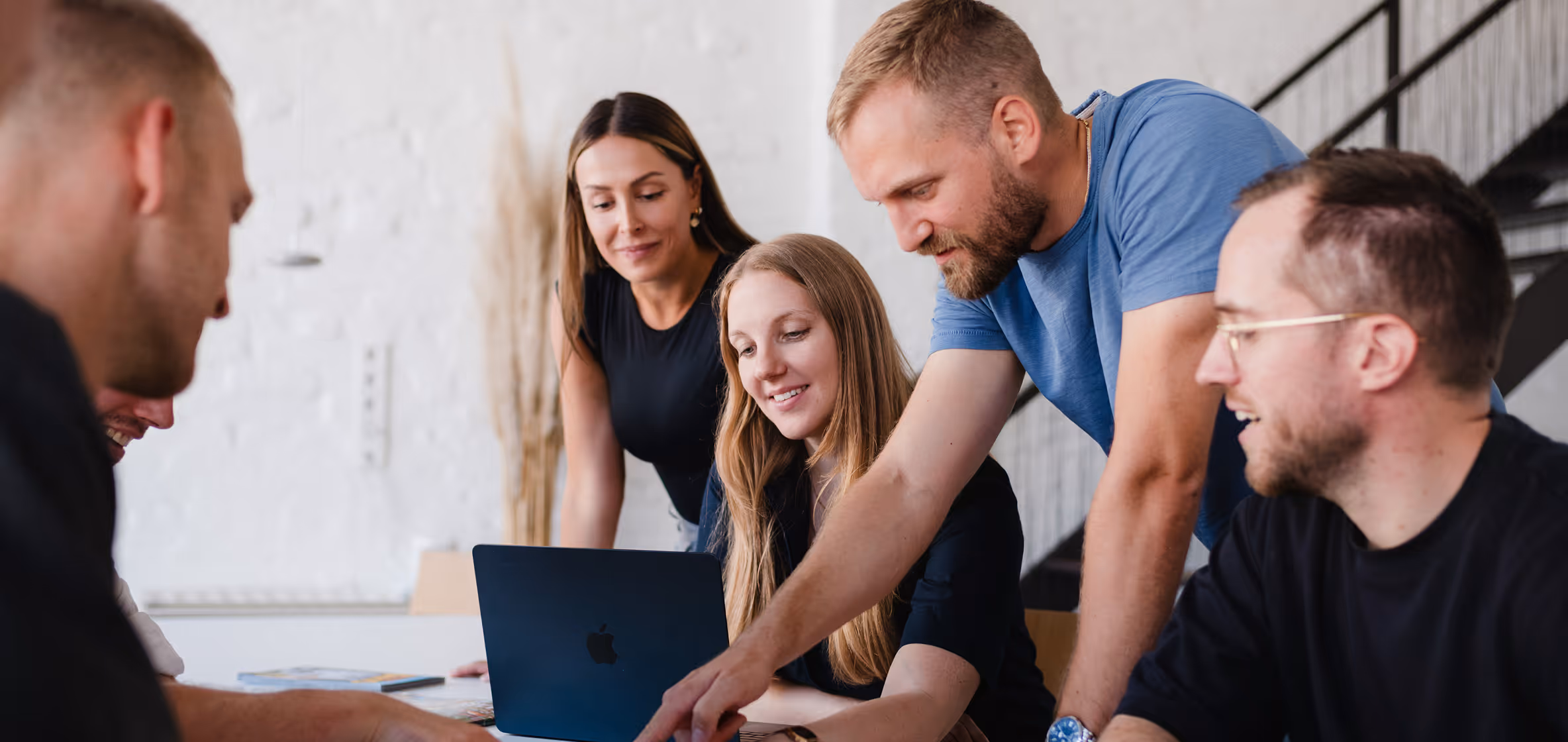 Fünf Personen arbeiten gemeinsam an einem Laptop in einem modernen Büro mit Treppe im Hintergrund.