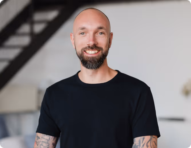 Lächelnder Mann mit Glatze, Bart und Tattoos an den Armen, trägt ein schwarzes T-Shirt vor einem Innenraum mit Treppe.