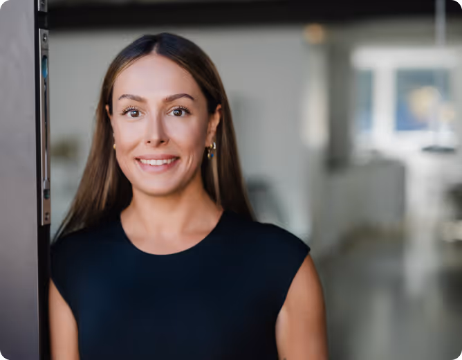 Junge Frau mit langen braunen Haaren und schwarzem ärmellosem Oberteil lächelt in einem modernen, hellen Innenraum.