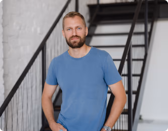Mann mit blauem T-Shirt und Bart steht vor einer Treppe mit schwarzem Geländer.
