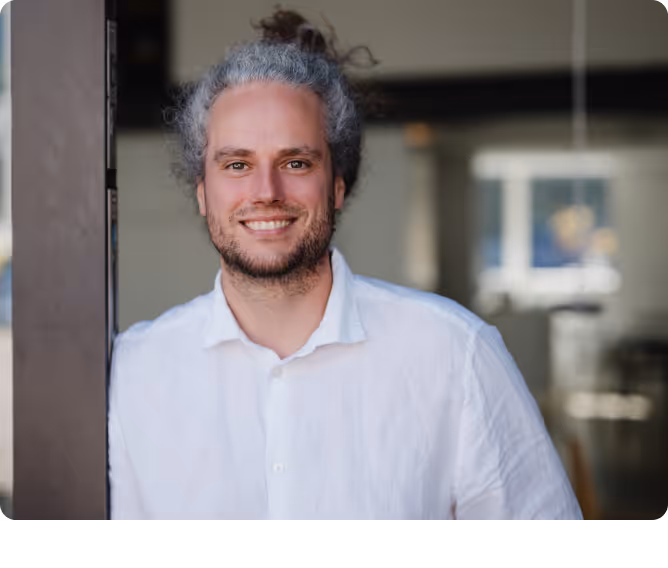 Mann mit lockigem grauem Haar und Bart trägt ein weißes Hemd und lächelt vor einem unscharfen Hintergrund.