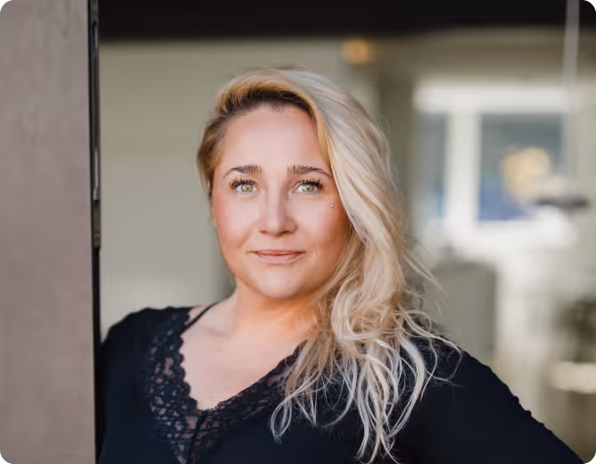 Porträt einer lächelnden Frau mit langen blonden Haaren, die vor einem unscharfen Hintergrund steht.