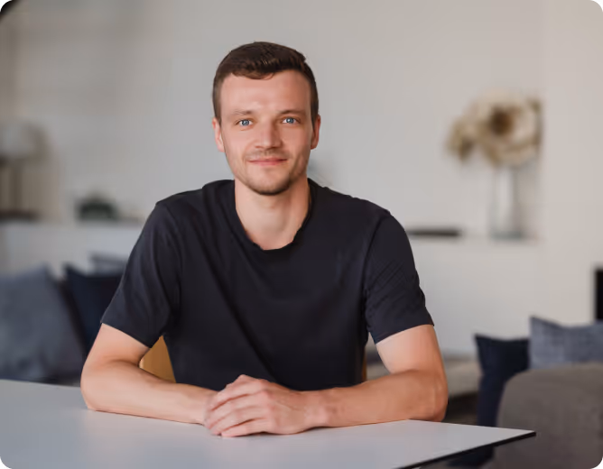 Junger Mann mit kurzem Haar und blauem T-Shirt sitzt lächelnd an einem Tisch vor verschwommenem Wohnzimmerhintergrund.