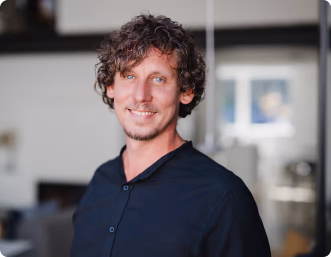 Ein lächelnder Mann mit lockigem braunem Haar und blauem Hemd vor einem unscharfen Büroraum.