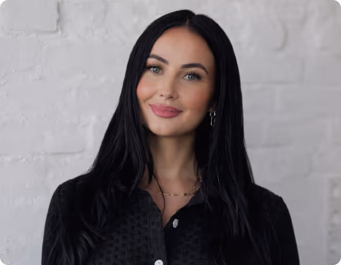 Porträt einer lächelnden Frau mit langen schwarzen Haaren und schwarzer Bluse vor weißer Steinmauer.