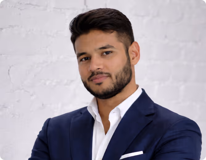 Junger Mann mit kurzem dunklem Haar und Bart in blauem Anzug vor weißer Ziegelwand.