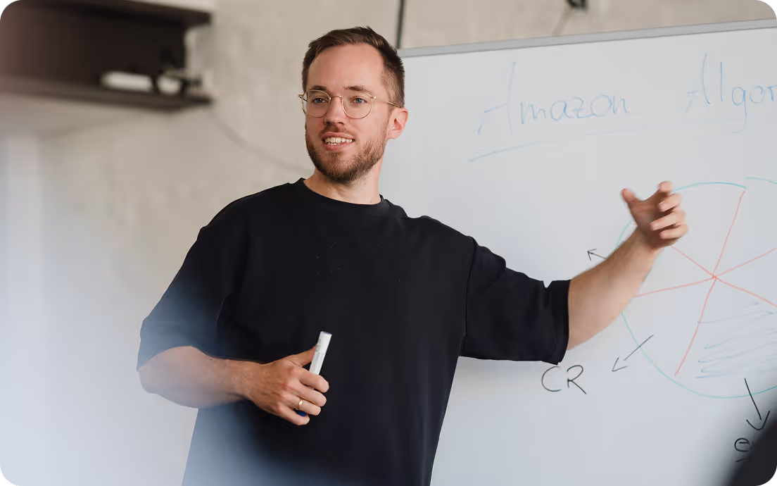 Ein Mann mit Brille und dunklem T-Shirt erläutert etwas vor einem Whiteboard mit Skizzen und dem Text "Amazon Algorithmus".
