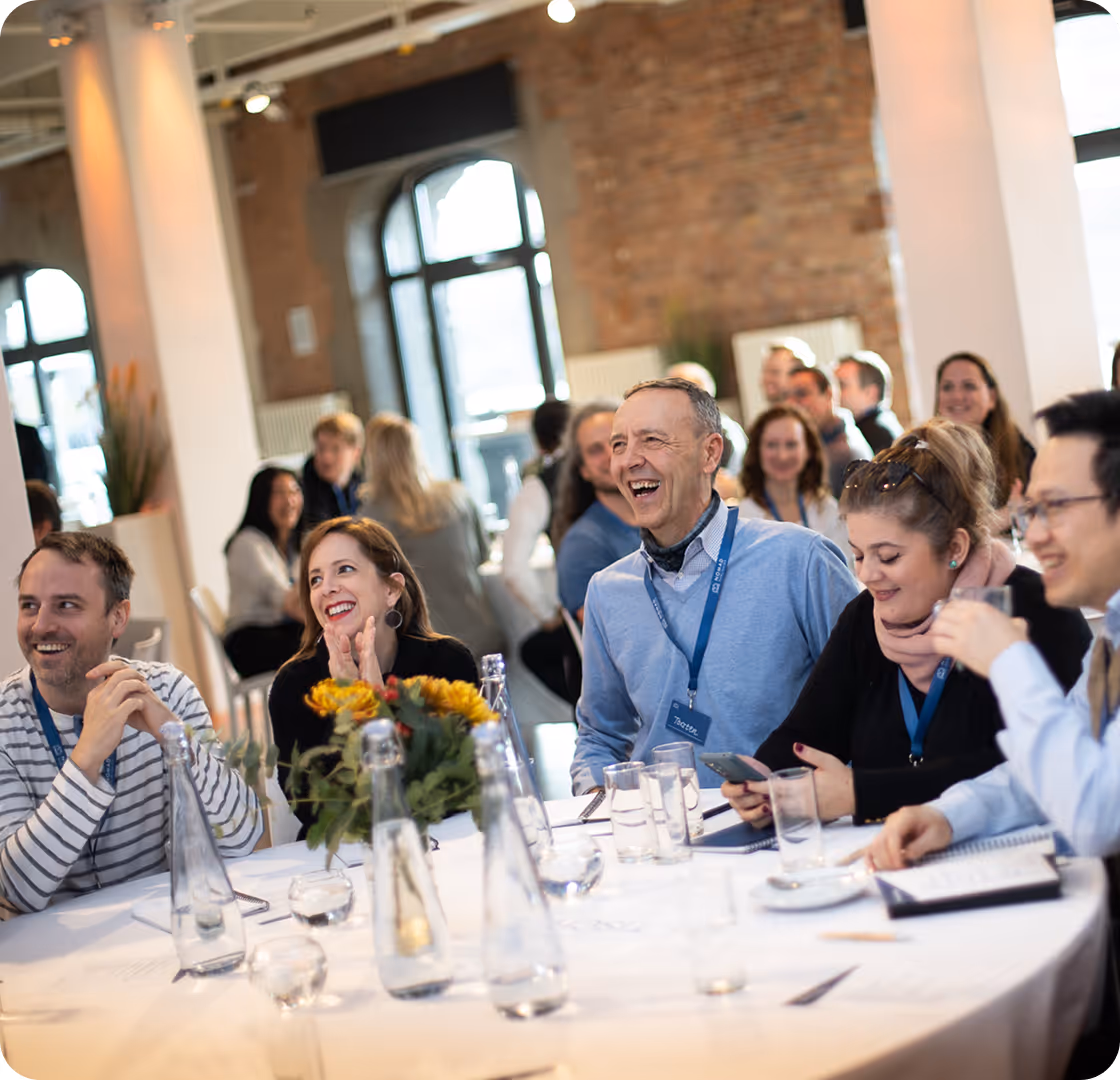 Menschen unterschiedlichen Alters und Geschlechts sitzen lachend an einem runden Tisch bei einer Veranstaltung oder einem Meeting.