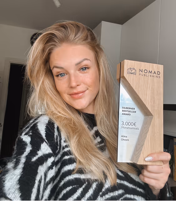 Eine junge Frau mit langen blonden Haaren hält einen hölzernen Award mit dem Schriftzug 'NOMAD PUBLISHING', 'SILBERNER BESTSELLER AWARD' und '3.000€ Monatsumsatz'.