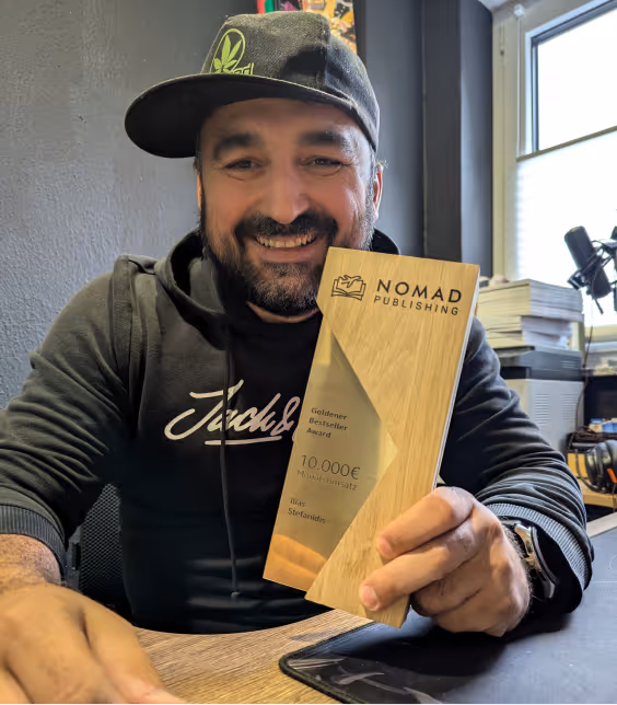 Mann sitzt an einem Tisch und hält einen hölzernen Nomad Publishing Award für 10.000 Euro, im Hintergrund Bücher und ein Fenster.