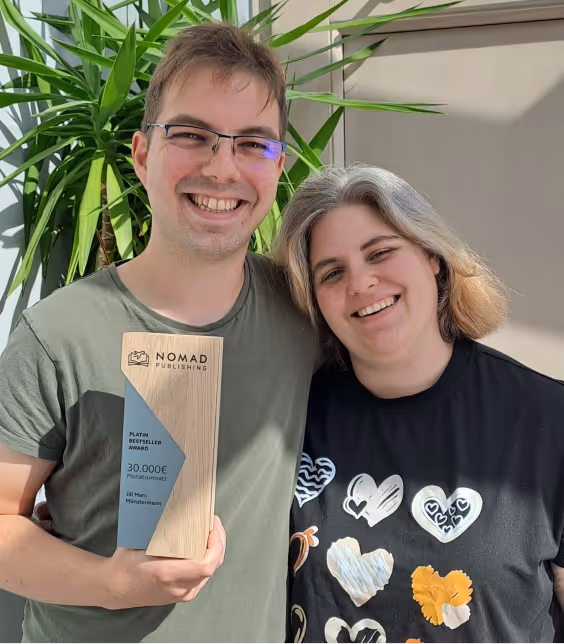 Zwei lächelnde Personen, eine hält einen hölzernen Nomad Publishing Platin Bestseller Award mit 30.000€ Preisgeld.