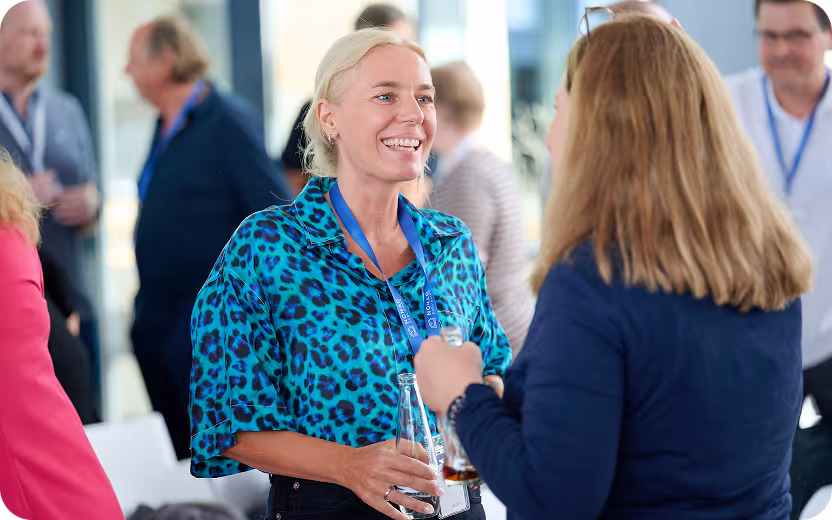 Eine lächelnde blonde Frau in einem blauen Leopardenmuster-Hemd spricht mit einer anderen Frau bei einer Veranstaltung.