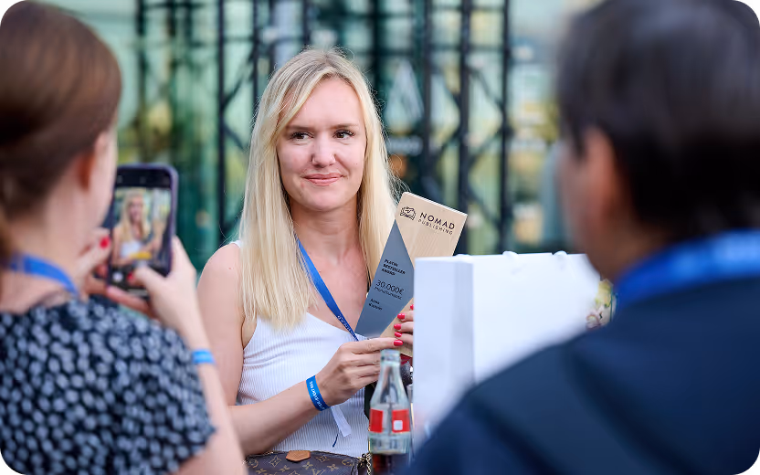 Blonde Frau mit weißem Top hält eine Auszeichnung von Nomad Publishing, während eine andere Person ein Foto davon macht.