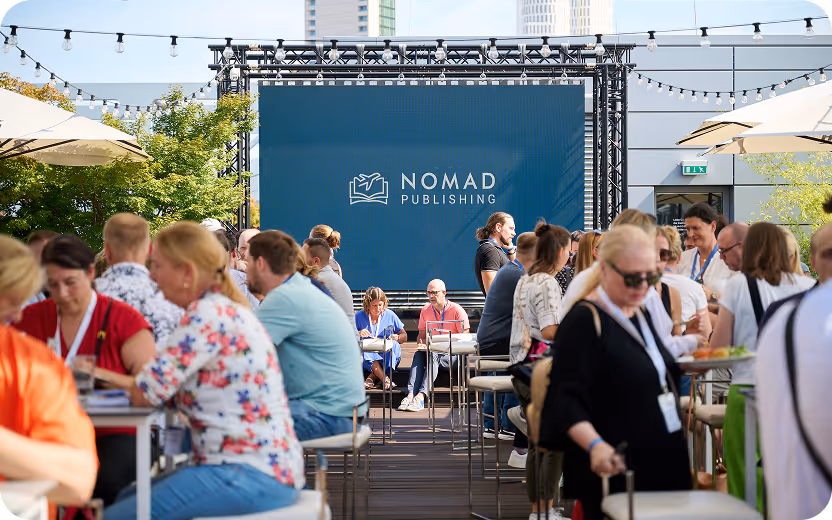 Menschen sitzen und unterhalten sich auf einer Terrasse mit einem großen Bildschirm, der das Logo von Nomad Publishing zeigt.