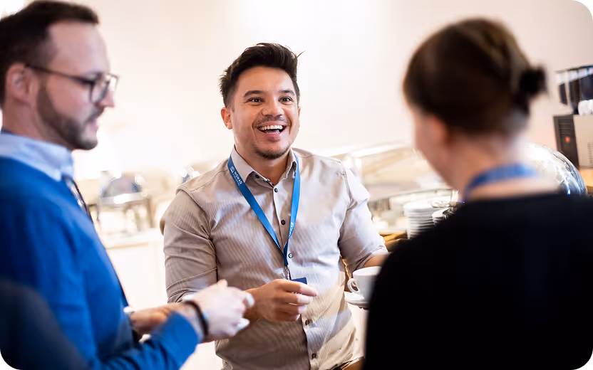 Drei Personen mit Namensschildern unterhalten sich lachend in einem hellen Raum während einer Kaffeepause.