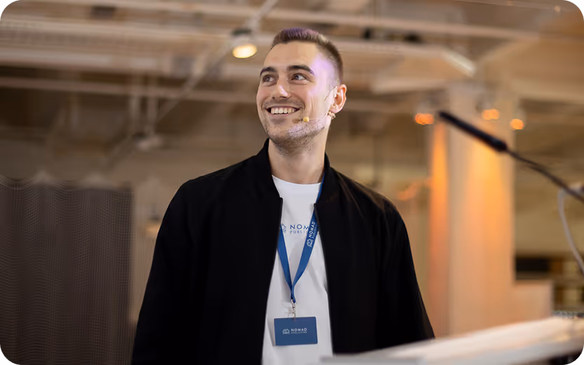 Lächelnder junger Mann mit Kurzhaarschnitt, der in einem modernen Raum mit Mikrofon und Namensschild spricht.