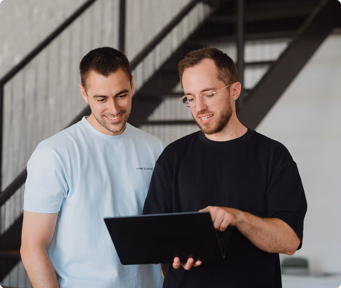 Zwei junge Männer stehen vor einer Treppe und schauen gemeinsam auf einen Laptop, einer zeigt auf den Bildschirm.