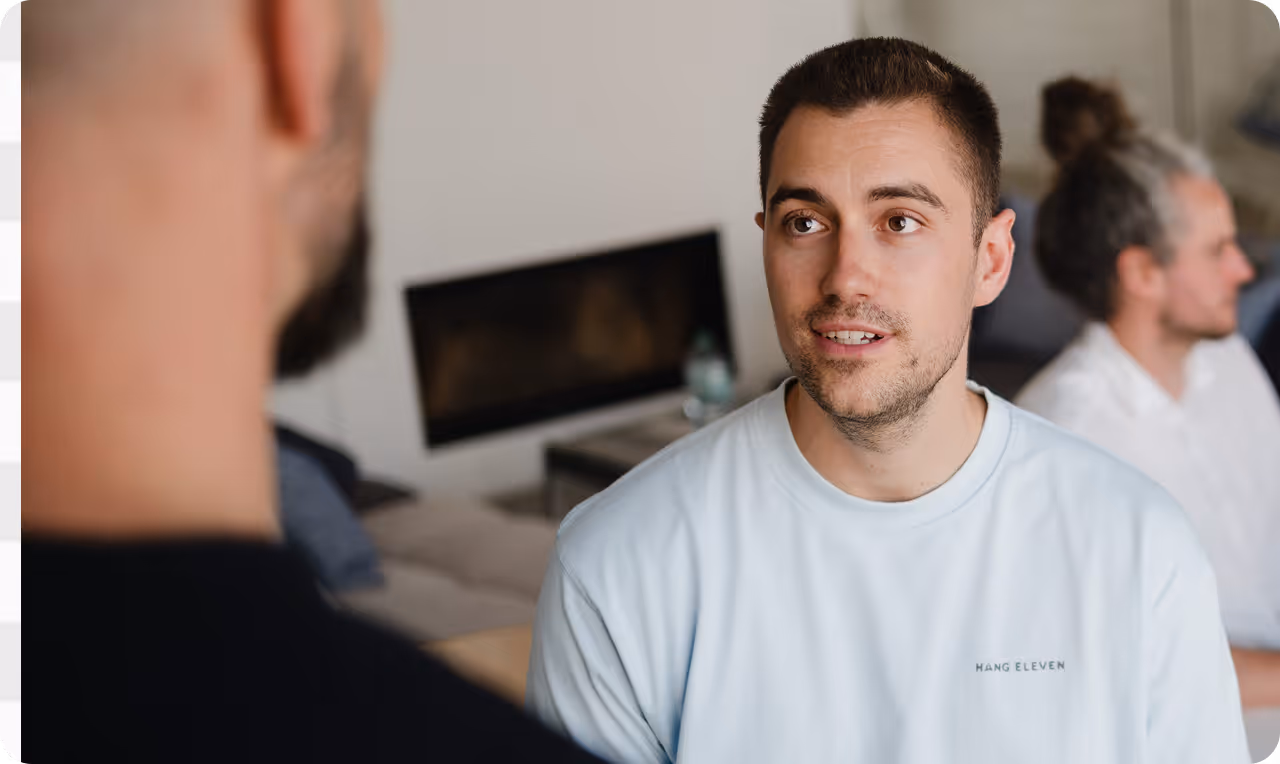 Junger Mann mit kurzem Haar und weißem T-Shirt, der einem anderen Mann zuhört, davor im unscharfen Vordergrund. Im Hintergrund sitzt ein Mann mit grauem Dutt.