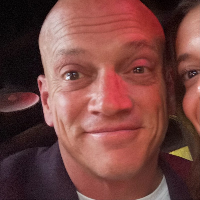 Close-up of a smiling bald man with a slight red light on his face, partially showing a woman on the right.