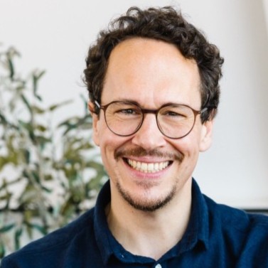 Smiling man with curly dark hair, glasses, and a short beard wearing a dark blue shirt in front of a blurred background with a plant.