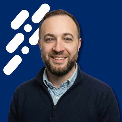 Smiling man with short hair and beard wearing a navy quarter-zip sweater over a light blue button-up shirt, set against a dark blue background with white abstract shapes.