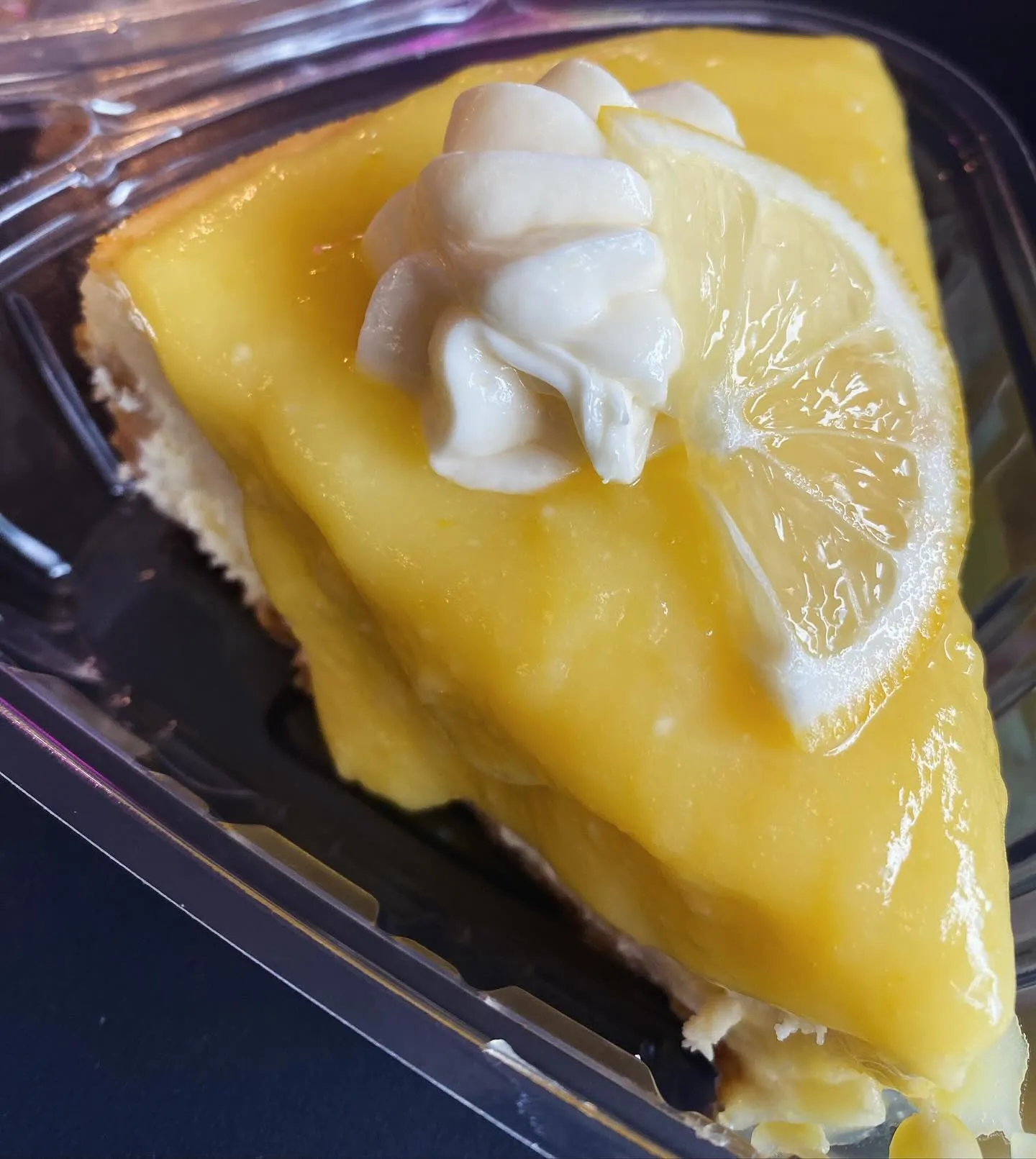A slice of lemon cheesecake displayed in a clear plastic container, showcasing its creamy texture and lemon topping.