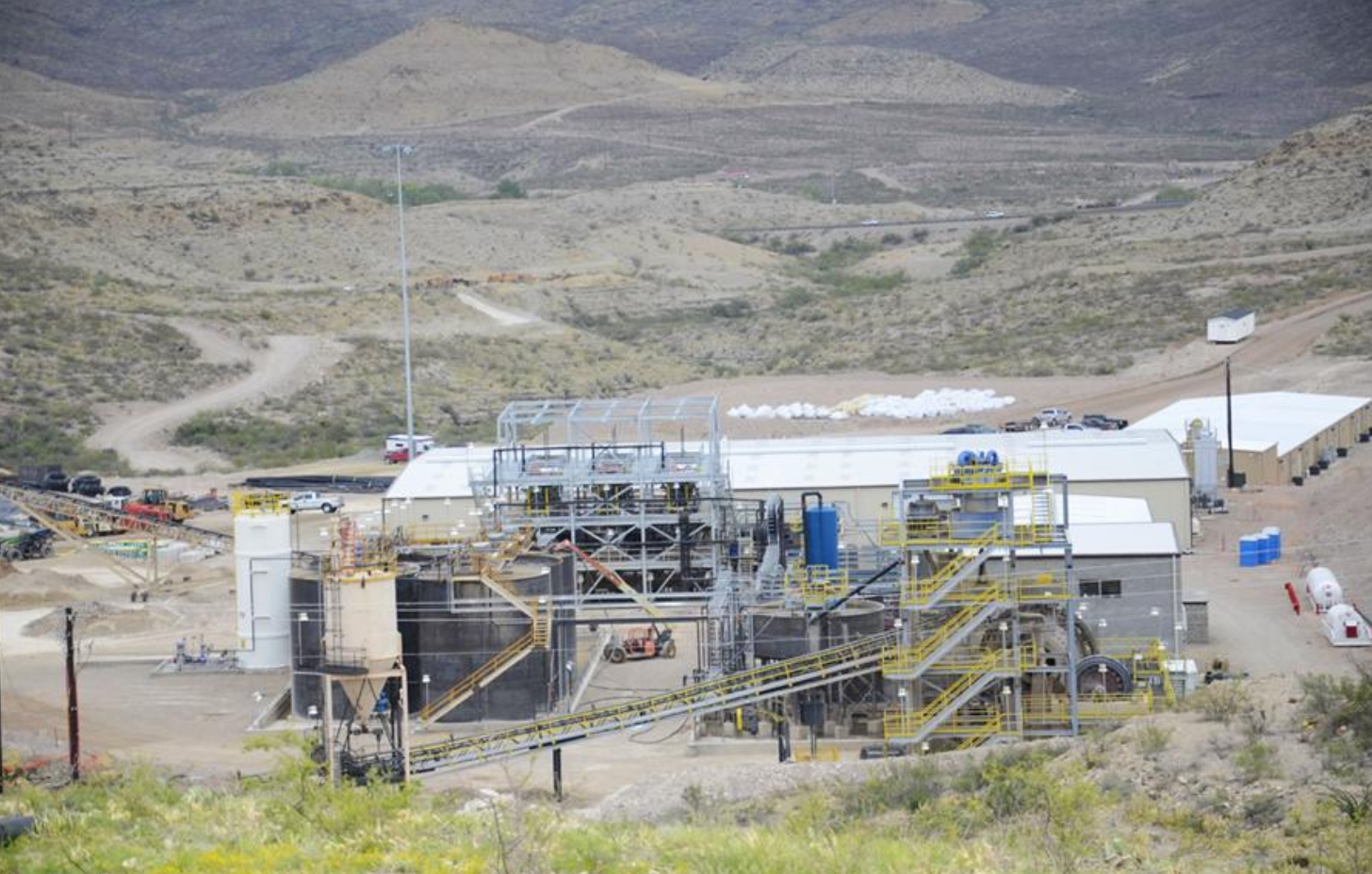Industrial mining facility with metal structures, conveyor belts, storage tanks, and buildings in a desert landscape.