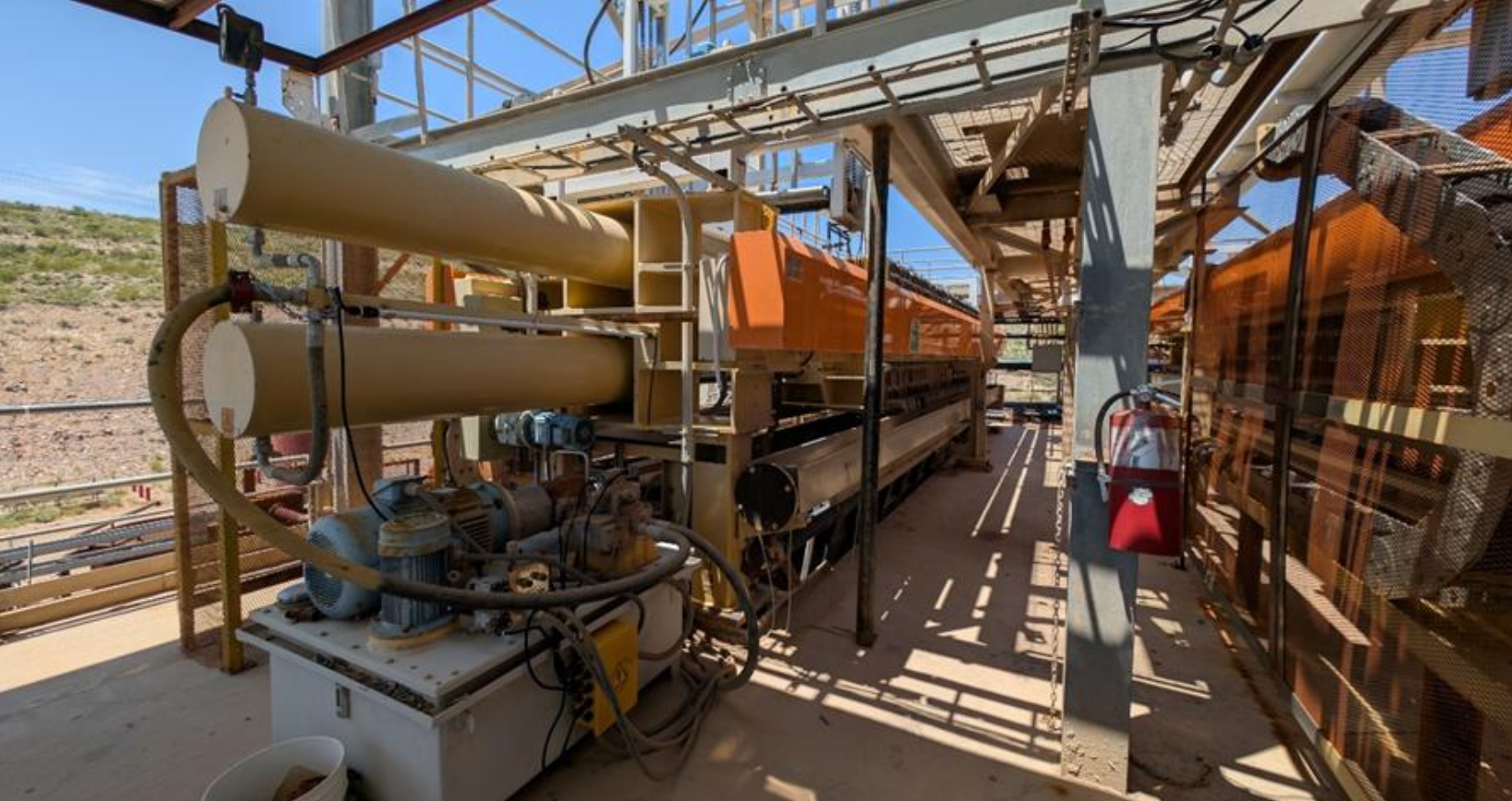 Industrial machinery with large yellow cylindrical components and an orange conveyor system under a metal framework.