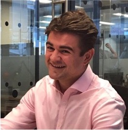 Man with short brown hair smiling and wearing a light pink button-up shirt in an office setting with glass walls.