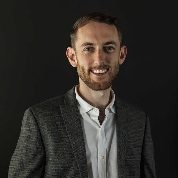 Smiling man with short brown hair and beard wearing a gray blazer and white shirt against a dark background.