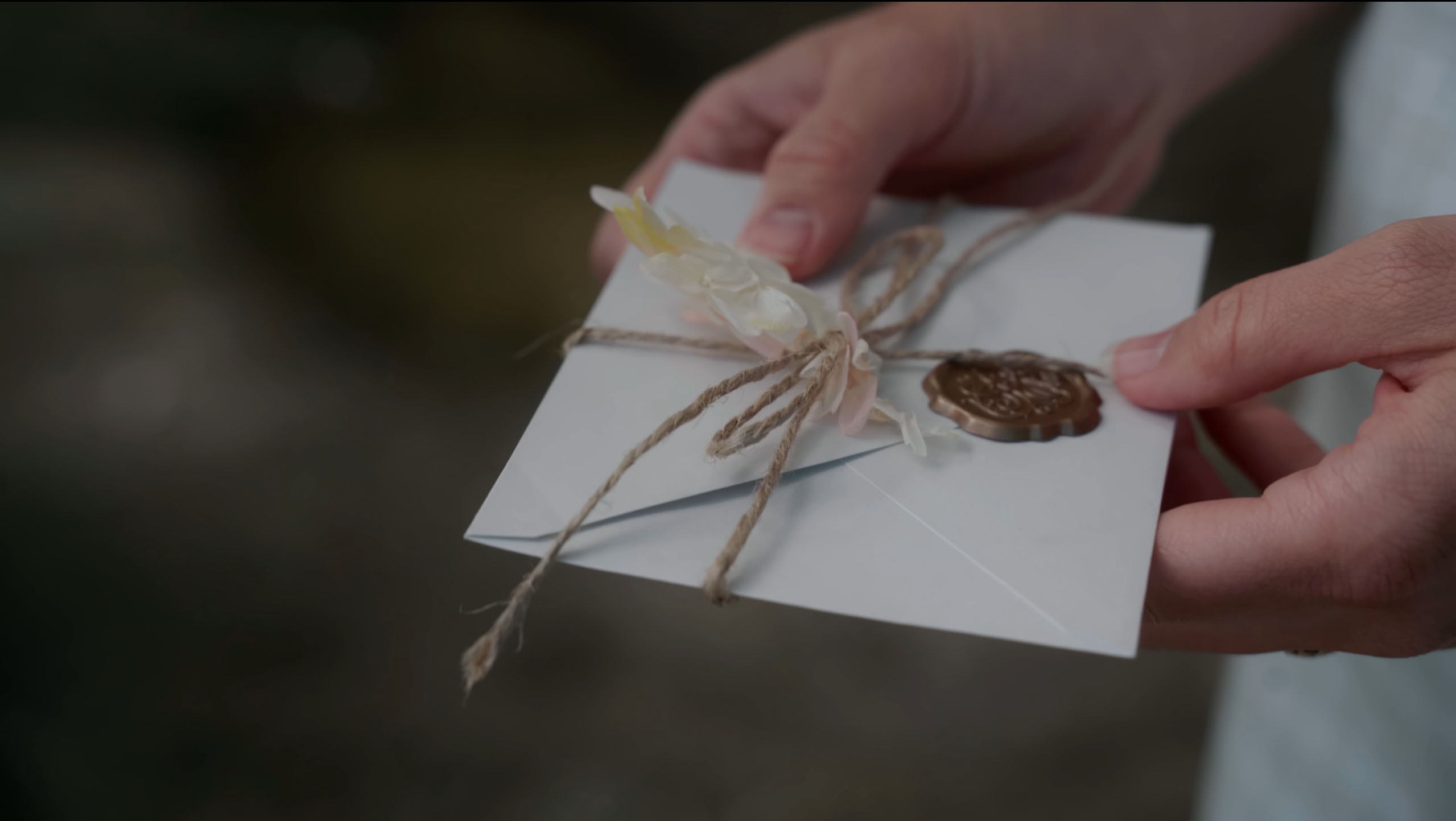 Close-up of hands holding a white envelope tied with twine, decorated with small flowers and a wax seal.