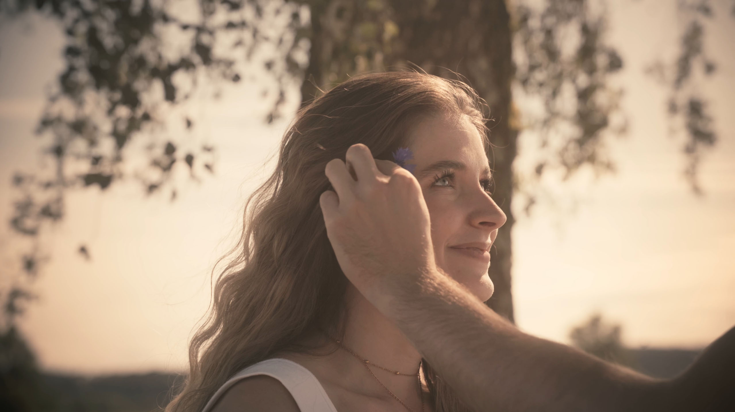 A woman smiles gently as a person places a small purple flower behind her ear outdoors near a tree.