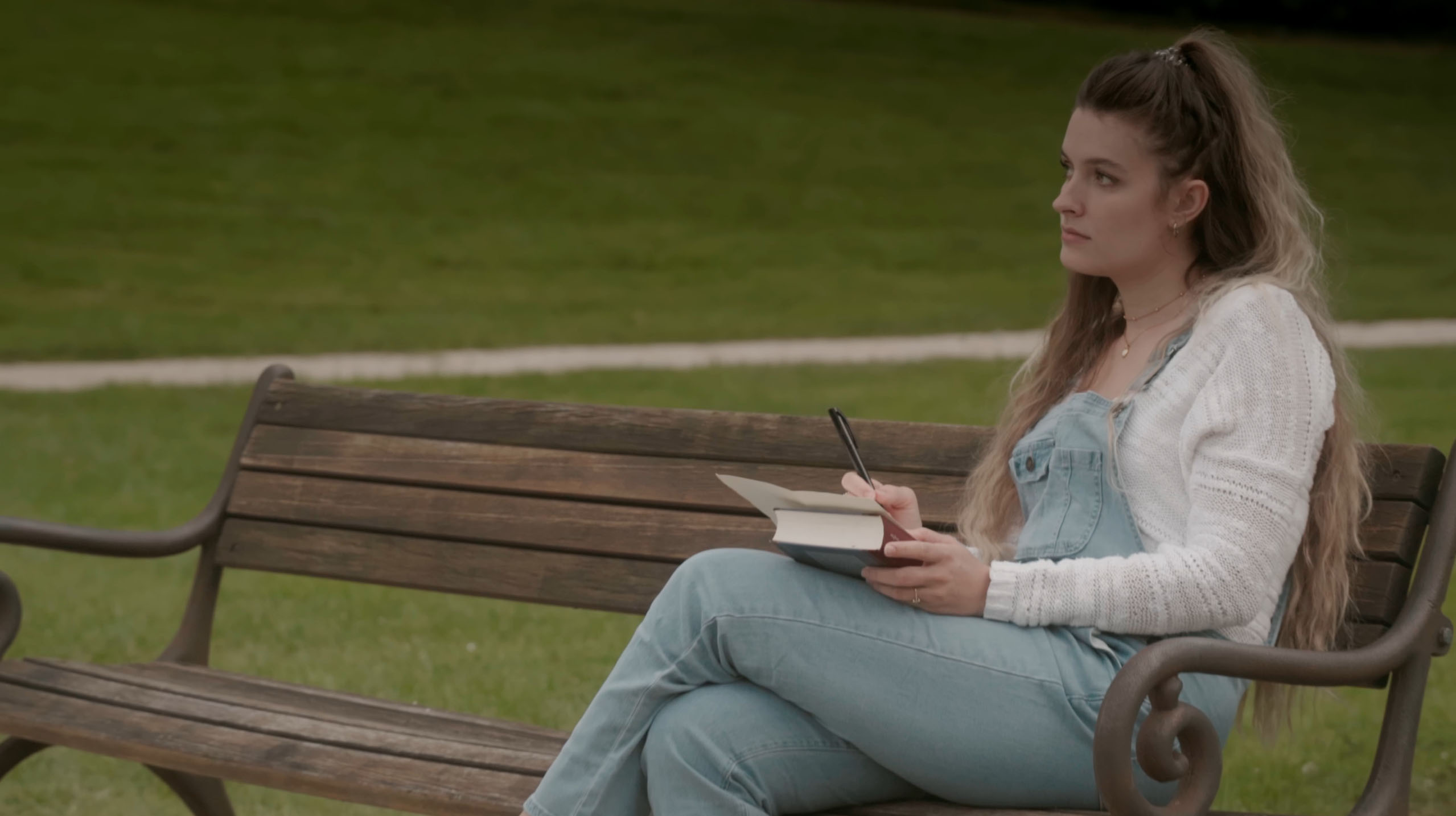 Young woman sitting on a wooden park bench writing in a notebook with a pen.