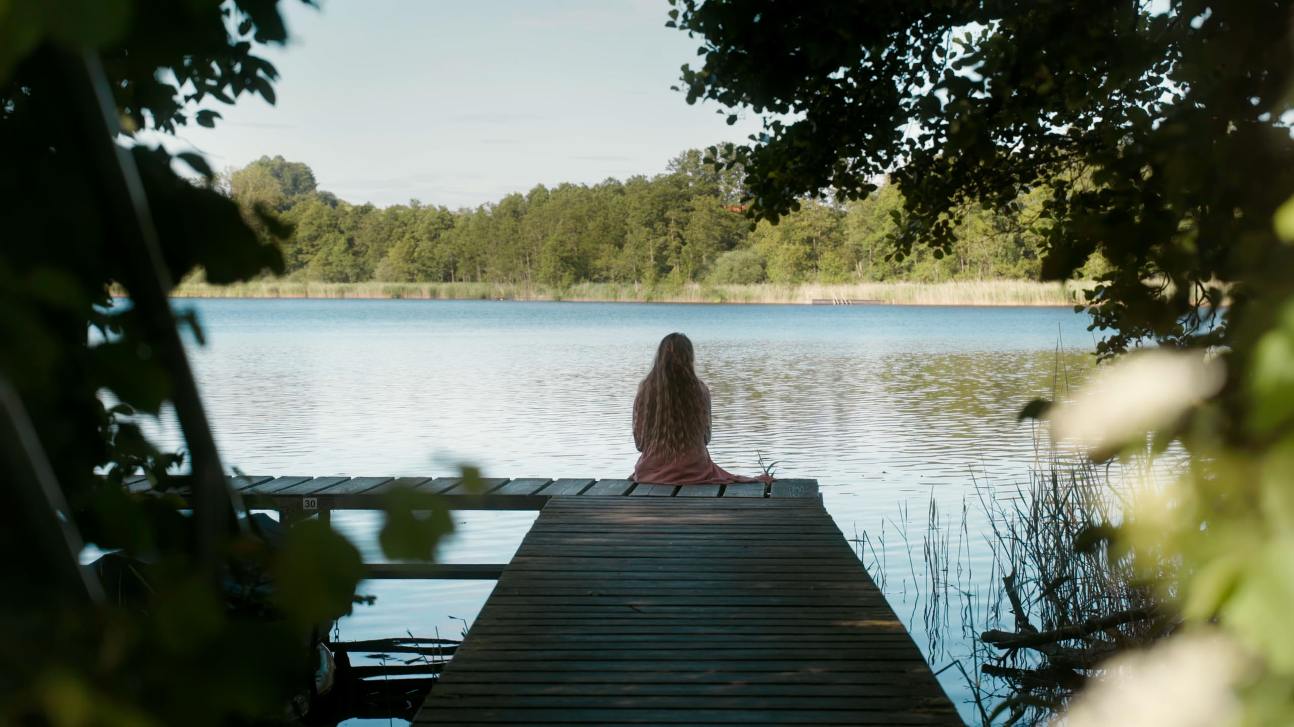 Person with long hair sitting alone on a wooden dock overlooking a calm lake surrounded by trees.