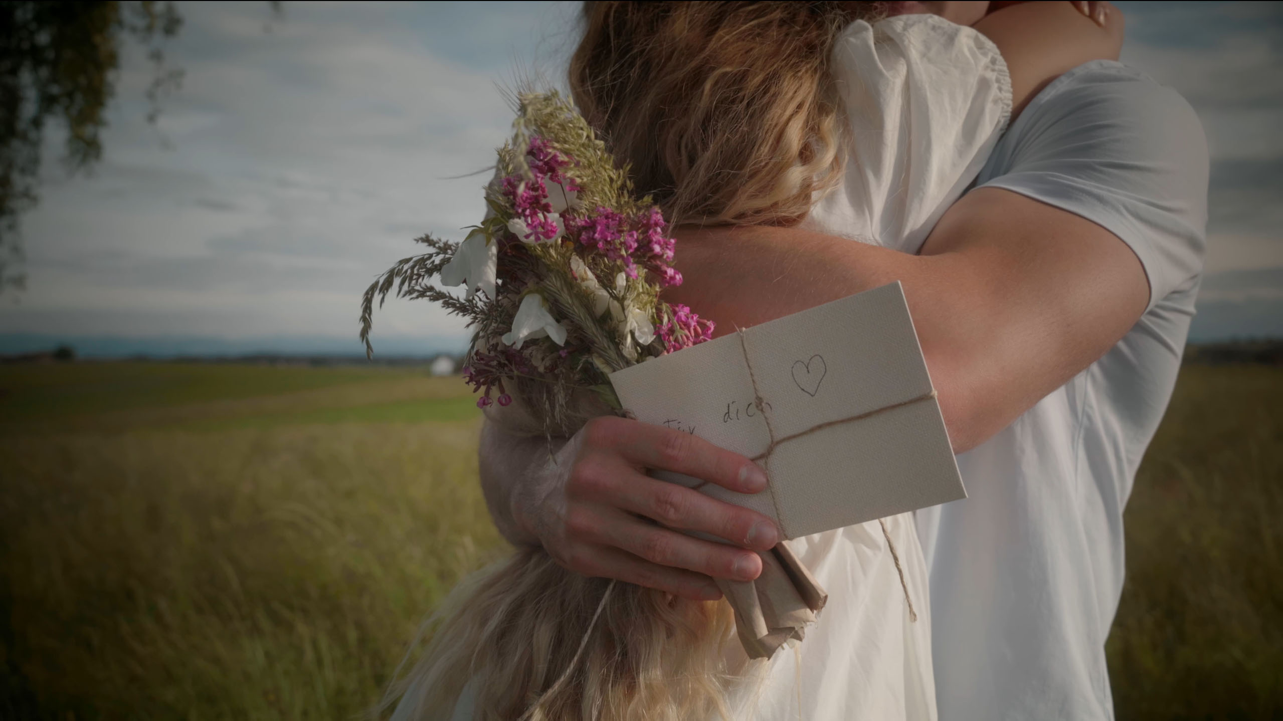 Couple embracing in a field, with one person holding a small bouquet of wildflowers and a tied envelope with a heart drawn on it.