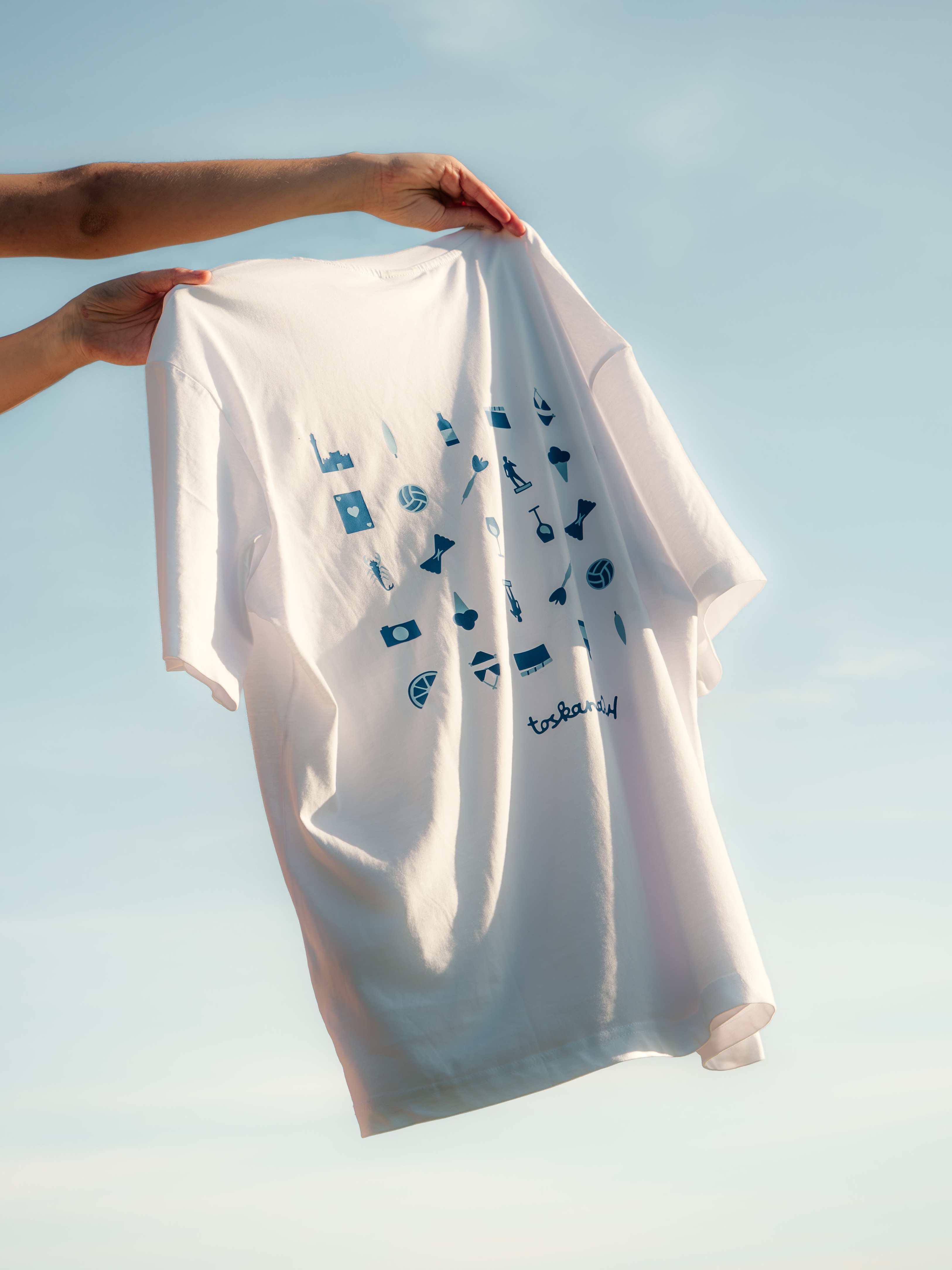 Person holding a white T-shirt with small blue icons and the word 'toskanaw' printed on it against a clear sky.