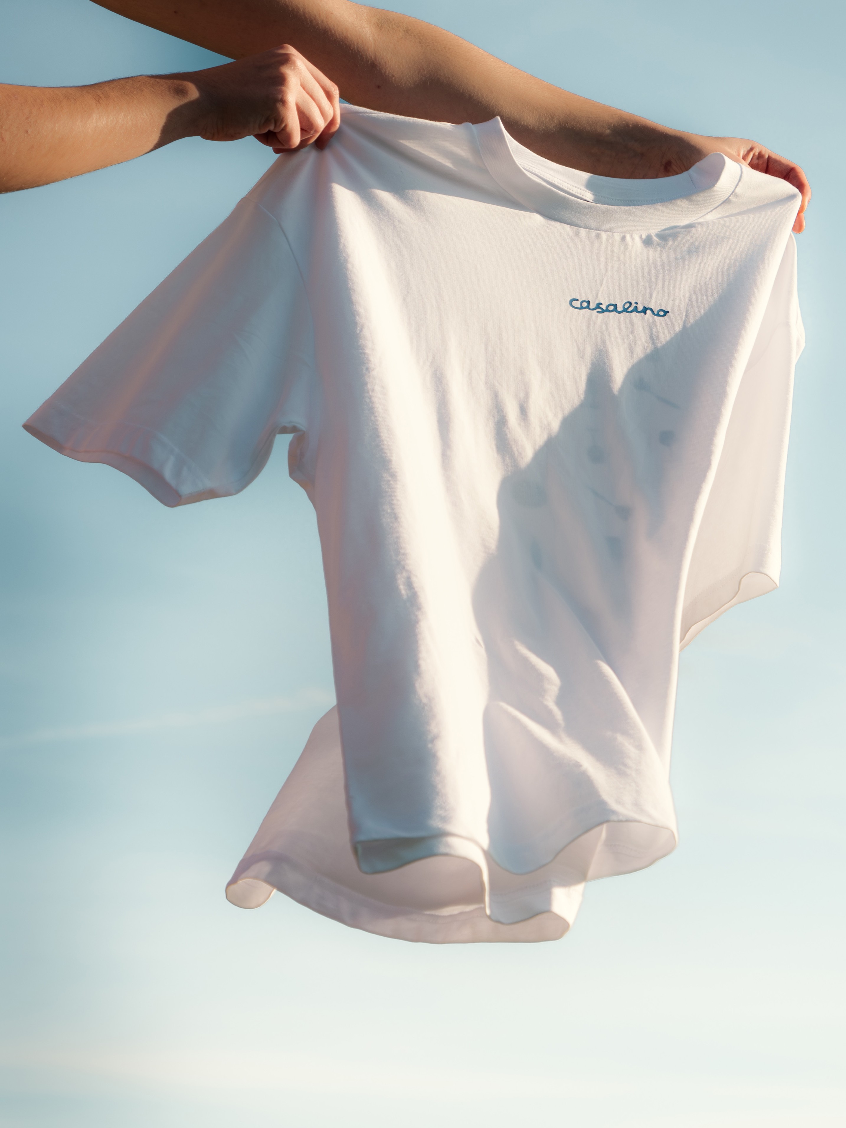 Hands holding a white T-shirt with the word 'casalino' printed near the collar against a clear blue sky.