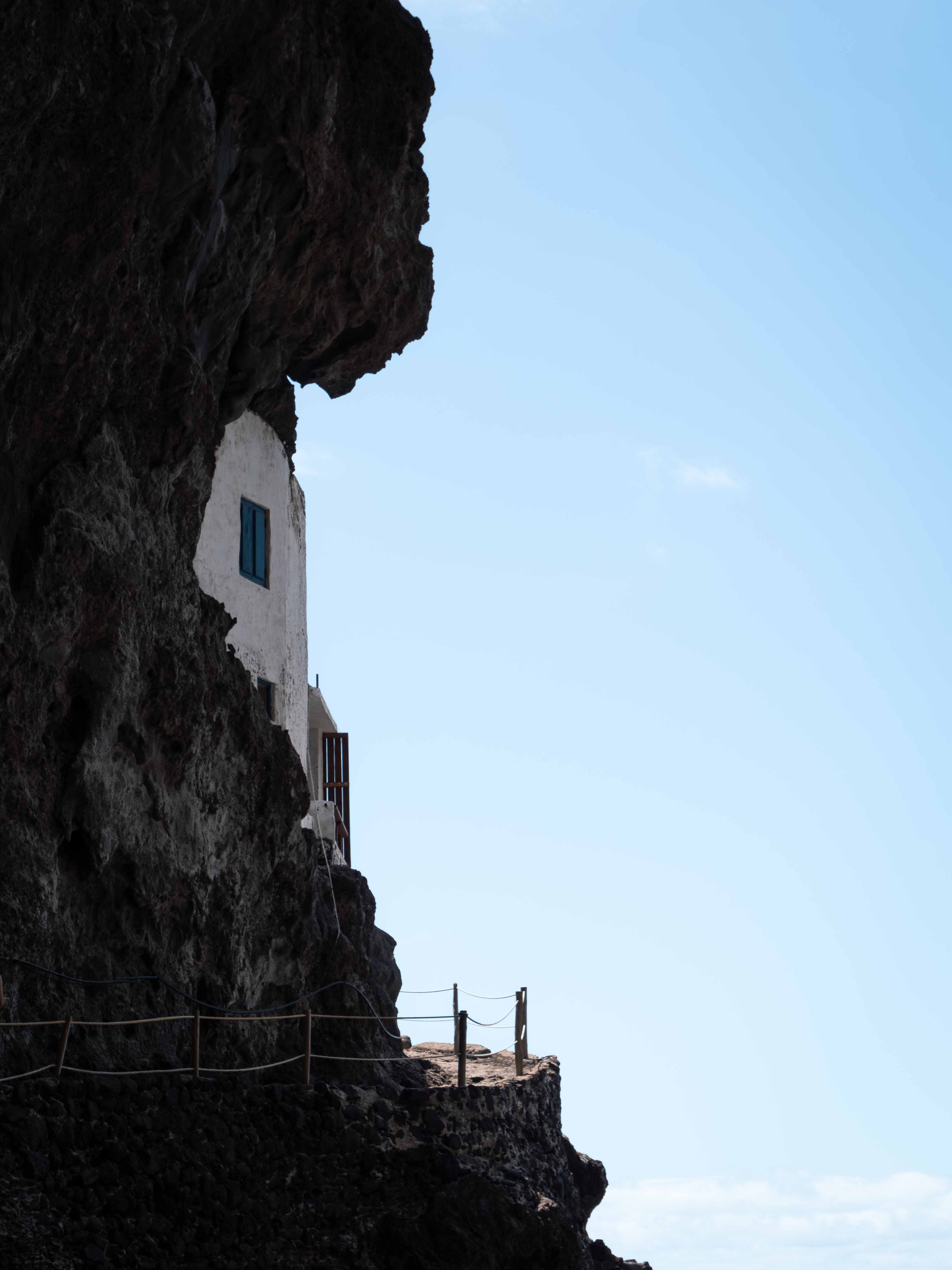 White building with blue window built into a dark rocky cliff against a clear blue sky.