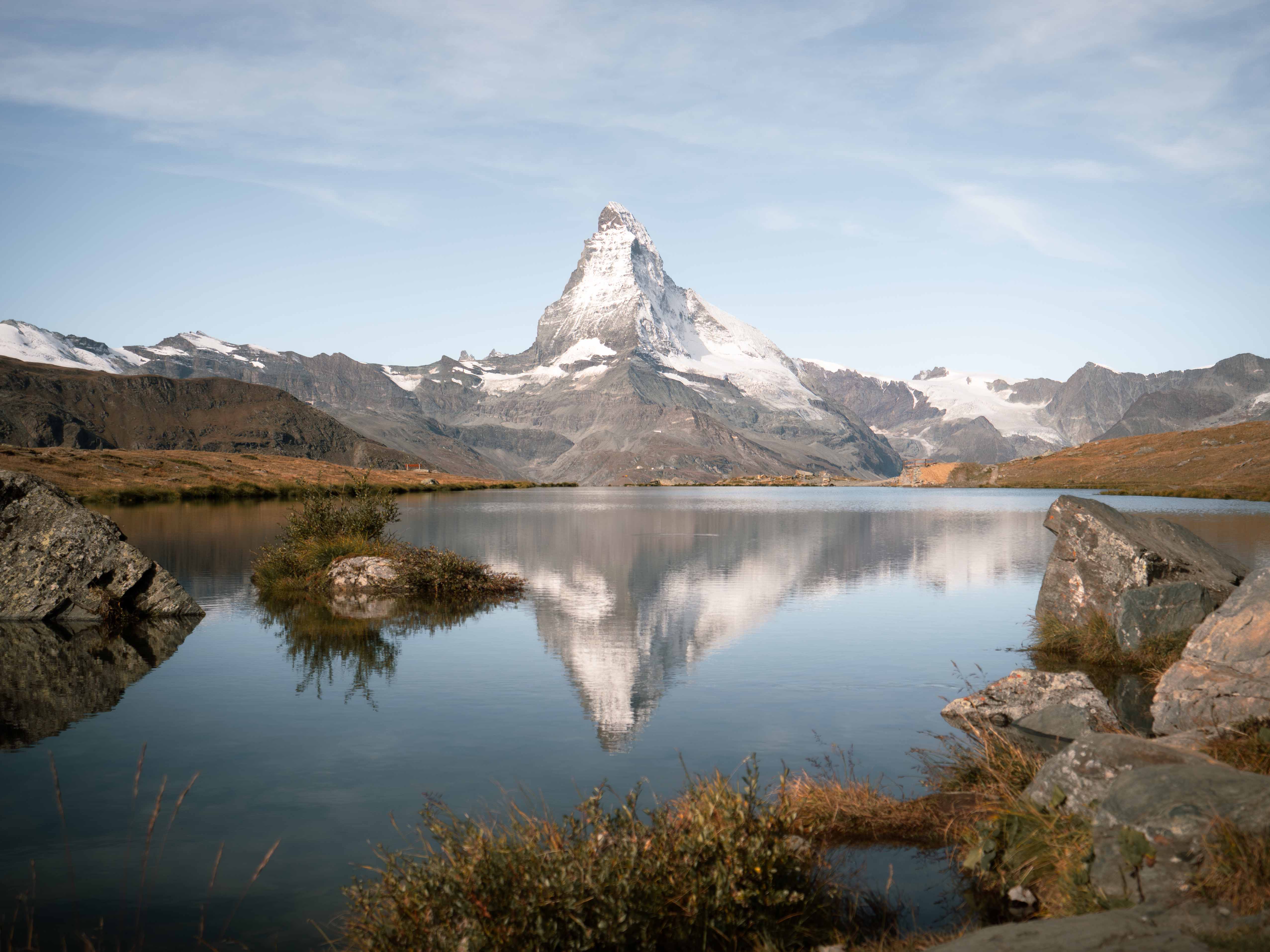 Snow-capped Matterhorn mountain reflected in a calm alpine lake surrounded by rocks and grass.