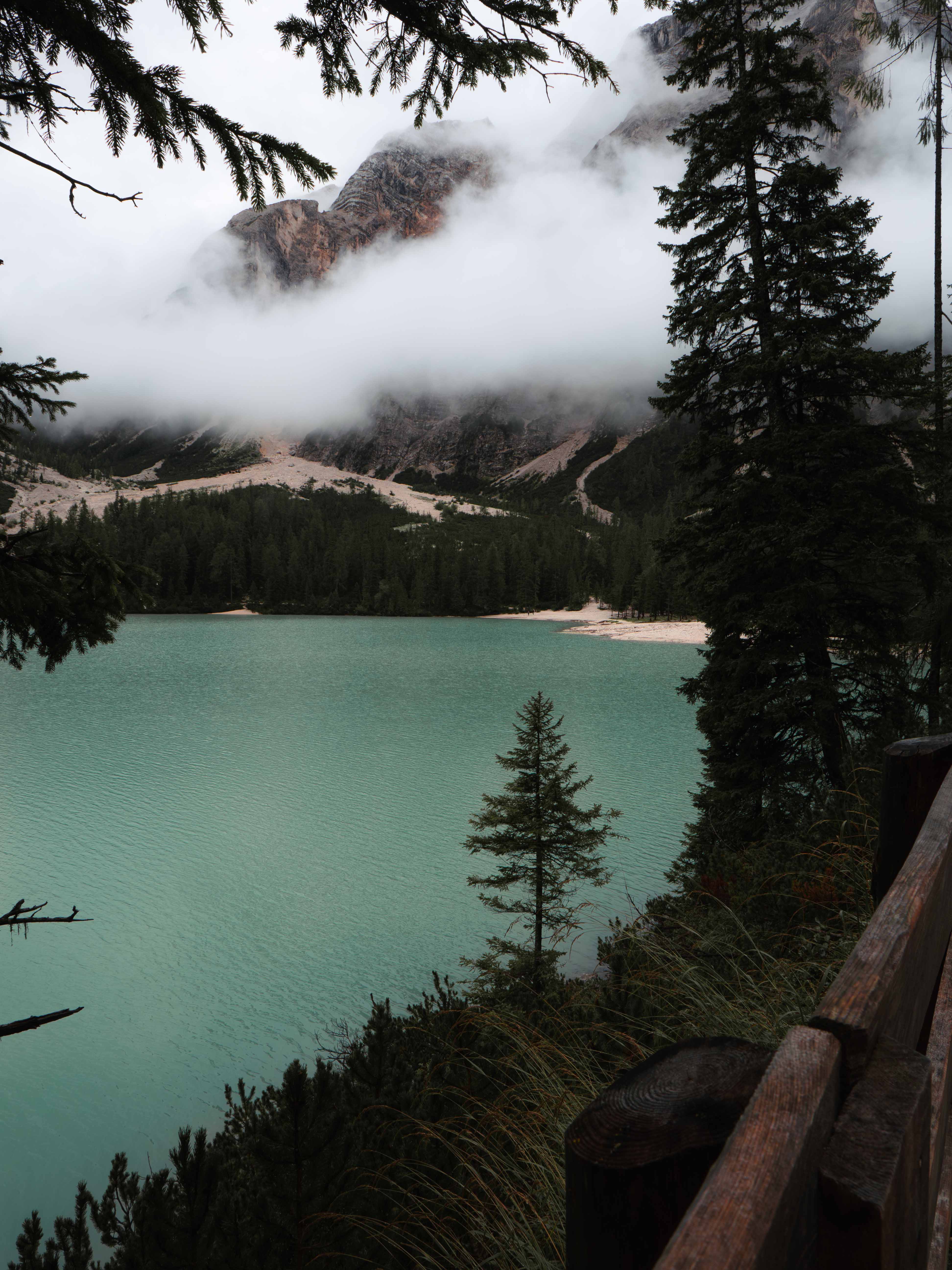 Calm turquoise lake surrounded by pine trees with mist-covered rocky mountains in the background.