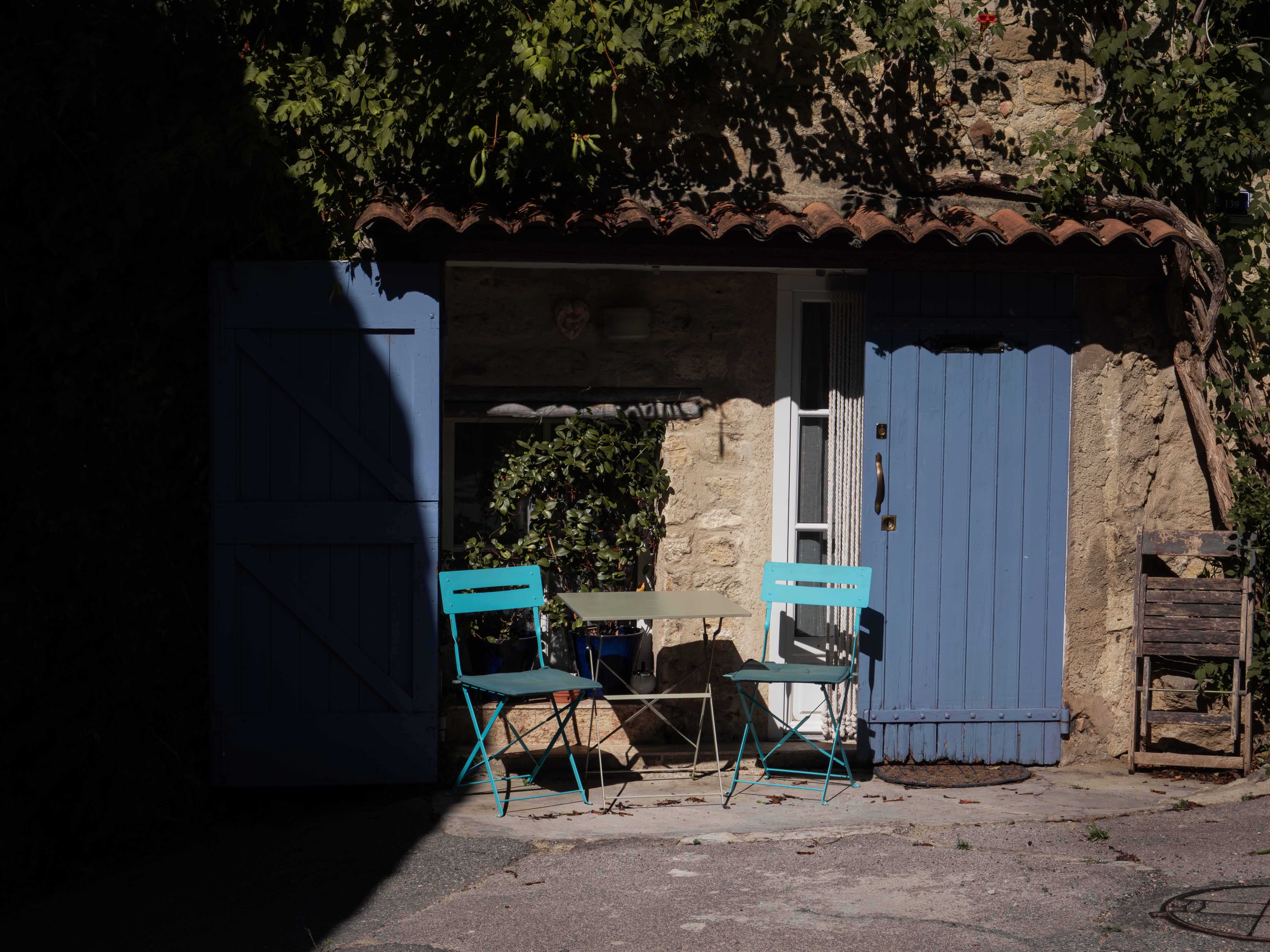 Small outdoor seating area with two turquoise metal chairs and a beige table in front of a stone wall with blue wooden doors and leafy vines.