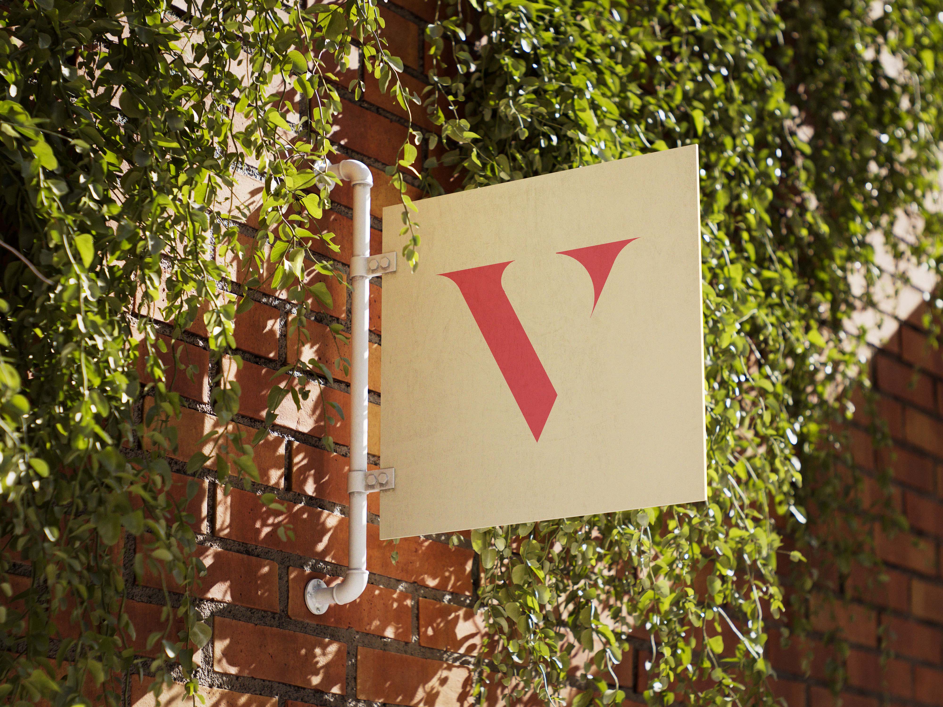 Square beige sign with a red stylized letter V mounted on a brick wall surrounded by hanging green foliage.