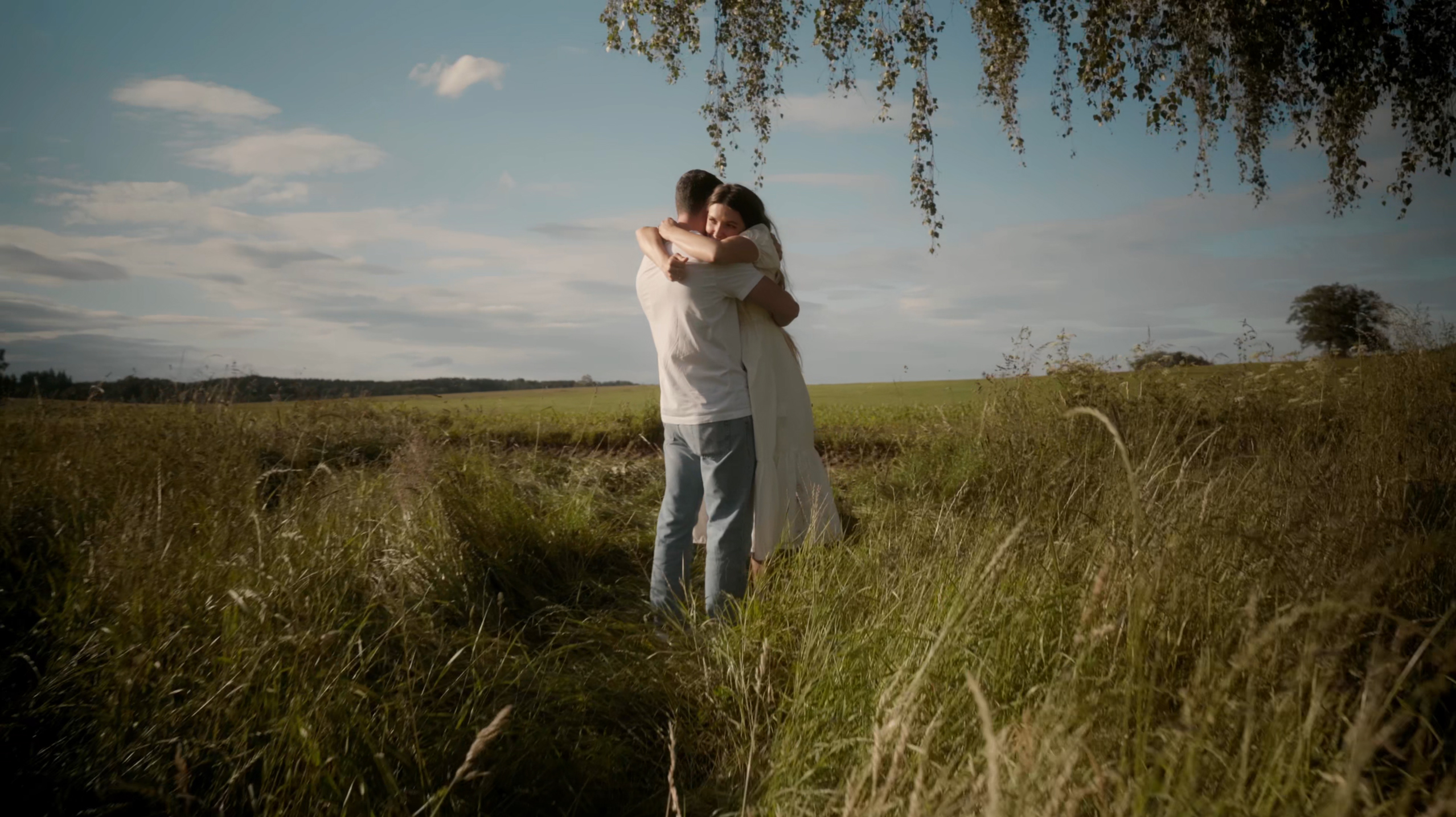 Couple hugging in a grassy field under a partly cloudy sky.