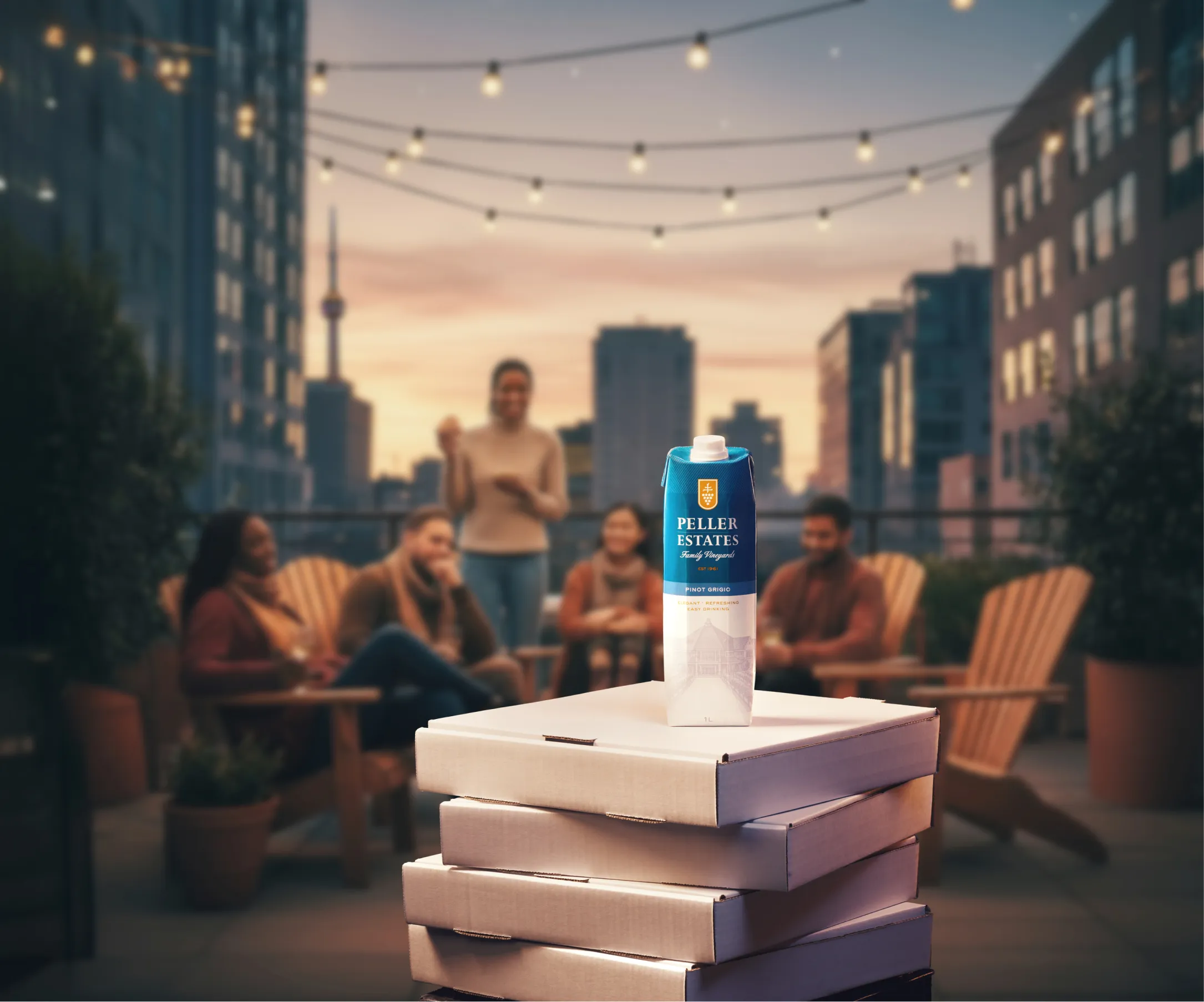 Peller Estate Wine in a 1 L carton sitting atop a stack of pizza boxes, with a rooftop patio party going on in the background.