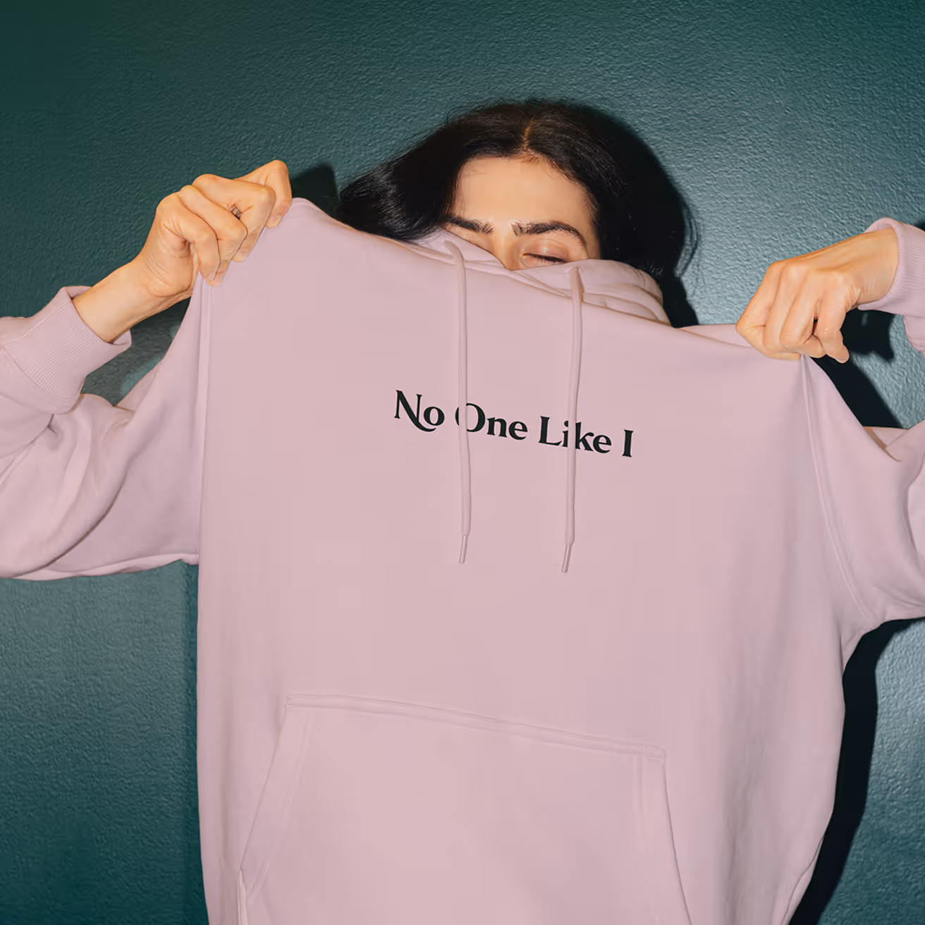Person pulling a light purple hoodie with text 'No One Like I' over their face against a dark green wall.